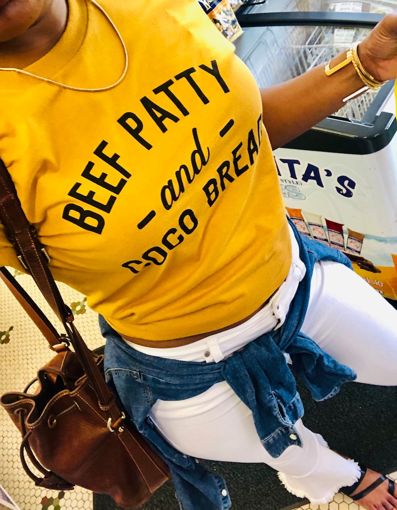 May include: A woman wearing a mustard yellow t-shirt with the text "Beef Patty - and Coco Bread" printed on it. She is also wearing white jeans and a denim jacket tied around her waist.