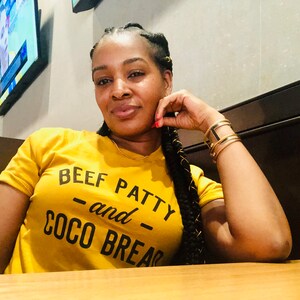 May include: A woman wearing a yellow t-shirt with the text "BEEF PATTY -and- COCO BREAD" in black lettering. She is sitting at a table with a wooden surface.