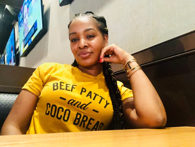 May include: A woman wearing a yellow t-shirt with the text "BEEF PATTY -and- COCO BREAD" in black lettering. She is sitting at a table with a wooden surface.