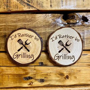 Może przedstawiać: Dwa drewniane podkładki z brązowym usłojeniem drewna. Każda podkładka zawiera tekst "I'd Rather Be Grilling" z grafiką łopatki i widelca.
