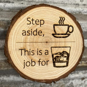 Puede incluir: Un posavasos redondo de madera con borde de corteza natural. El posavasos está grabado con las palabras "Step aside, This is a job for" encima de una taza de café y un vaso de whisky con hielo.