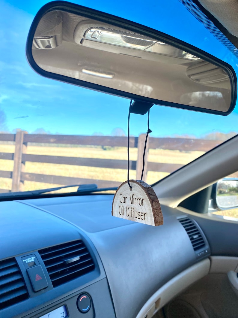 Peut inclure: Un diffuseur d'huile pour r&eacute;troviseur de voiture en bois suspendu au r&eacute;troviseur d'une voiture. Le diffuseur a la forme d'un demi-cercle et porte la mention "Car Mirror Oil Diffuser" grav&eacute;e dessus.