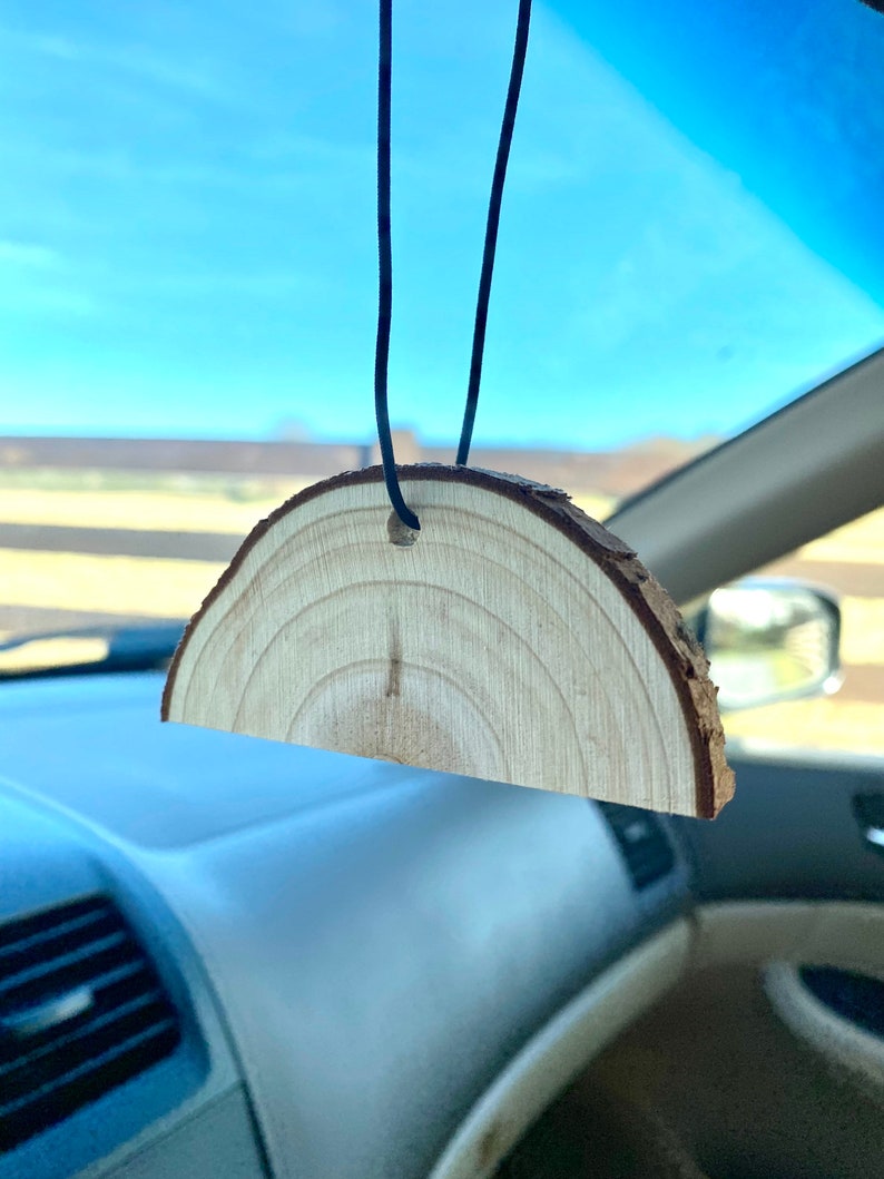 Peut inclure: Un diffuseur d'air de voiture en bois en forme de demi-cercle, suspendu &agrave; une ficelle noire. Le bois est brun clair avec un grain visible.