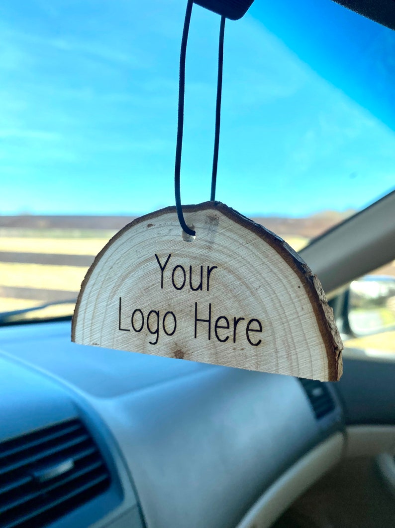 Peut inclure: Un diffuseur d'air de voiture en bois avec le texte "Your Logo Here" grav&eacute; dessus. Le diffuseur est suspendu &agrave; un r&eacute;troviseur.