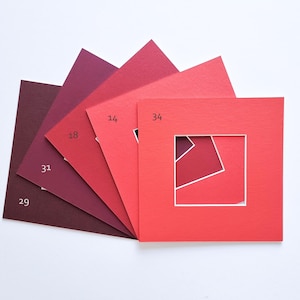 Puede incluir: Un abanico de cinco paspart&uacute;s cuadrados de papel en varios tonos de rojo, desde el burdeos intenso hasta el coral brillante. Cada paspart&uacute; presenta un recorte cuadrado central y est&aacute; etiquetado con los n&uacute;meros 29, 31, 18, 14 y 34.