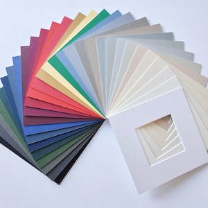 Puede incluir: Una colecci&oacute;n en abanico de cartones para paspart&uacute; cuadrados dispuestos en un espectro de colores, que van desde el negro profundo, azul marino y verde bosque hasta el rojo vibrante, dorado y varios tonos de gris, crema y blanco, con un corte central.