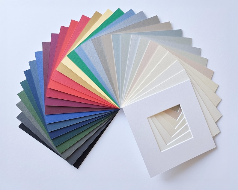 Puede incluir: Una colecci&oacute;n en abanico de cartones para paspart&uacute; cuadrados dispuestos en un espectro de colores, que van desde el negro profundo, azul marino y verde bosque hasta el rojo vibrante, dorado y varios tonos de gris, crema y blanco, con un corte central.