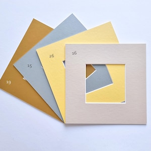 Puede incluir: Un conjunto de cuatro paspart&uacute;s cuadrados de papel dispuestos en abanico. Los colores incluyen amarillo mostaza, gris claro, amarillo p&aacute;lido y beige. Cada paspart&uacute; presenta un recorte cuadrado en el centro y est&aacute; etiquetado con los n&uacute;meros 19, 15, 26 y 16.