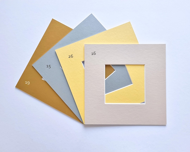 Puede incluir: Un conjunto de cuatro paspart&uacute;s cuadrados de papel dispuestos en abanico. Los colores incluyen amarillo mostaza, gris claro, amarillo p&aacute;lido y beige. Cada paspart&uacute; presenta un recorte cuadrado en el centro y est&aacute; etiquetado con los n&uacute;meros 19, 15, 26 y 16.