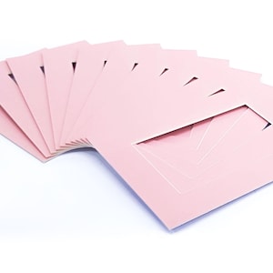 May include: A collection of pale pink picture mounts, each with a rectangular aperture and a beveled edge. The mounts are fanned out, showing the cut-out shape. The background is white.
