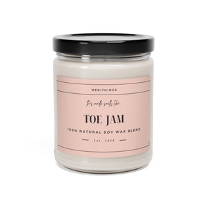 May include: A glass jar candle with a pink label that reads "MEDITHINGS This candle smells like TOE JAM 100% NATURAL SOY WAX BLEND Est. 2018".