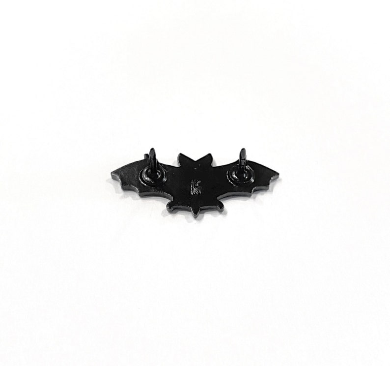 May include: Black metal bat pin with a star design on the back. The pin has two metal posts on the back for attaching to clothing.
