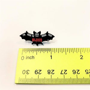 May include: Black enamel pin shaped like a bat with the text "I'm here to draw your blood" in white and red lettering. The bat has two googly eyes. The pin is measured against a ruler, showing it is approximately 1 inch wide.