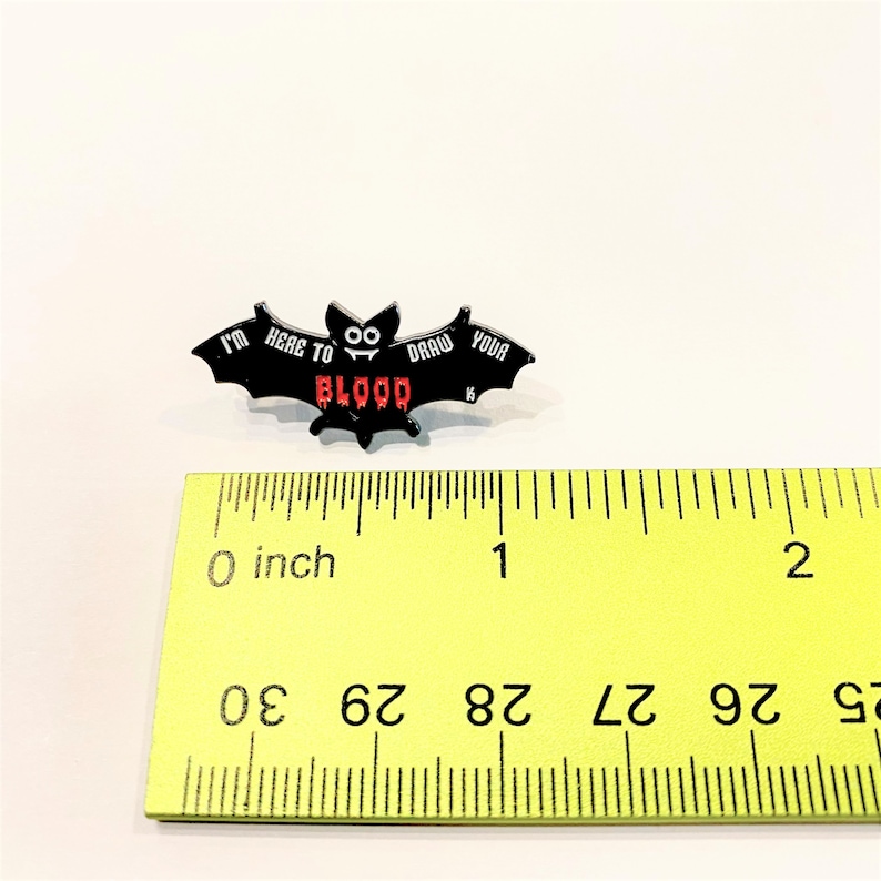 May include: Black enamel pin shaped like a bat with the text "I'm here to draw your blood" in white and red lettering. The bat has two googly eyes. The pin is measured against a ruler, showing it is approximately 1 inch wide.