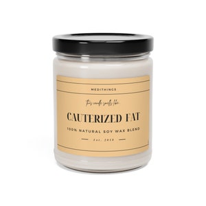 May include: A glass jar candle with a black lid and a beige label. The label reads "MEDITHINGS", "This candle smells like", "CAUTERIZED FAT", "100% NATURAL SOY WAX BLEND", and "Est. 2018".