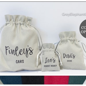 May include: Three beige drawstring bags of different sizes. The largest bag says "Finley's CARS", the medium bag says "Dad's COINS", and the smallest says "Leo's POCKET MONEY". The image also shows six colour options.