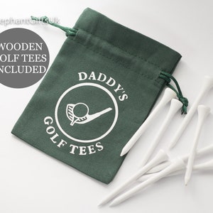 May include: A dark green drawstring bag printed with "DADDY'S GOLF TEES" and a golf ball graphic. Several white wooden golf tees are scattered around the bag. The text "WOODEN GOLF TEES INCLUDED" is also visible.