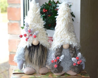 Könnte beinhalten: Zwei graue und weiße Gnome mit langen weißen Fellmützen und roten Beerenakzenten. Die Gnome stehen auf einer Holzkiste mit einem grün-braunen Muster.