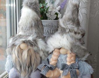 May include: Two plush gnomes with faux fur hats and beards. One gnome has a long, flowing beard, while the other has braided hair and bows. The gnomes are in shades of grey, white, and blue, with wooden noses.