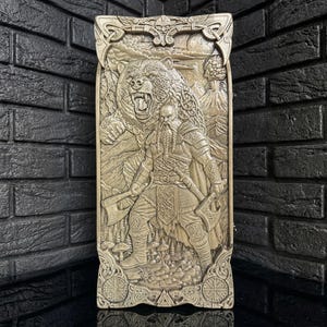 May include: A beige rectangular relief carving depicting a Viking warrior with axes facing a roaring bear. The carving has intricate details of the warrior's clothing, the bear's fur, and the surrounding landscape. The top and bottom of the carving have decorative borders.