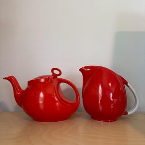 May include: A bright red ceramic teapot with a circular handle on the lid and a matching red creamer pitcher featuring a white handle. Both pieces have a smooth, glossy finish and rounded, bulbous shapes, showcasing a vibrant solid red color.