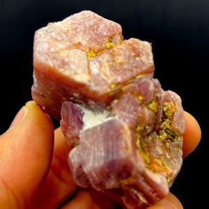 May include: A cluster of pink ruby crystals with a rough, natural texture. The crystals are intergrown and have a slightly metallic sheen.