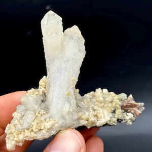 May include: A cluster of clear quartz crystals, featuring two main points. The crystals are a pale, translucent white, set on a base of smaller, beige crystals and a dark matrix. The structure is rough and natural.
