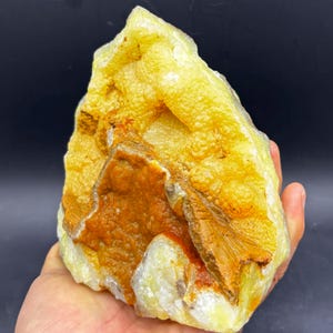 May include: A large, rough-hewn mineral specimen with a mix of yellow, orange, and white hues. The surface has a textured, bubbly appearance, with areas of crystalline structure. The rock is held in a hand, showcasing its size and natural beauty.