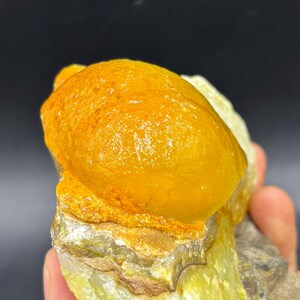 Könnte beinhalten: Nahaufnahme einer Mineralprobe mit einer abgerundeten, goldgelben Formation auf einer rauen, strukturierten Basis. Die Basis zeigt Grün-, Gelb- und Brauntöne, mit einem dunklen Hintergrund.