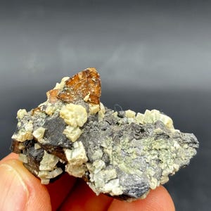 May include: A natural mineral specimen with a dark grey matrix, speckled with light green and off-white crystals. A prominent, angular, orange-brown crystal formation is at the top. The specimen is held in a hand against a dark background.
