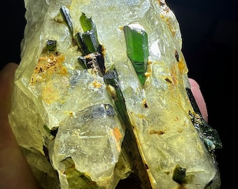 May include: A cluster of clear quartz crystals with embedded green tourmaline crystals. The quartz crystals are translucent and have a rough, natural texture. The tourmaline crystals are dark green and have a smooth, glassy surface.