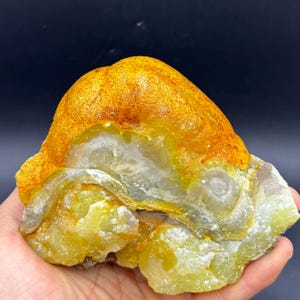 May include: A raw, natural gemstone with a layered appearance. The top portion is a textured, golden-orange color, transitioning to a pale yellow and white base. The stone is held in a hand, showcasing its unique mineral composition.