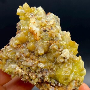 May include: A close-up of a mineral specimen featuring a cluster of yellow-green, crystal formations. The crystals have a radiating, needle-like structure, with hints of brown and white. The background is dark.