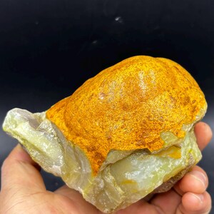 Könnte beinhalten: Ein rauer, ungeschliffener Edelstein mit einem geschichteten Aussehen. Die obere Schicht ist ein leuchtendes, strukturiertes Orange, während die Basis hellgrün ist. Der Stein wird in einer Hand gehalten und zeigt seine natürliche, rohe Schönheit. Der Hintergrund ist schwarz.