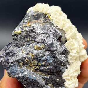 May include: A close-up of a mineral specimen featuring a dark, metallic gray base with patches of golden pyrite crystals. A cluster of creamy white, layered crystals sits atop the gray mineral, creating a striking contrast. The specimen is set against a dark background.