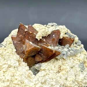 May include: A close-up of a mineral specimen with reddish-brown, pyramidal crystals. The crystals are set against an off-white and grey matrix. The rough texture of the mineral creates a natural appearance. The crystals are a striking feature.