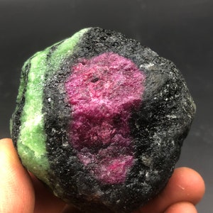 Puede incluir: Una gema de rubí zoisita en bruto y sin cortar. La piedra presenta un centro magenta vibrante rodeado por una matriz negra. Zoisita verde es visible en un lado. Esta piedra natural es un espécimen único.