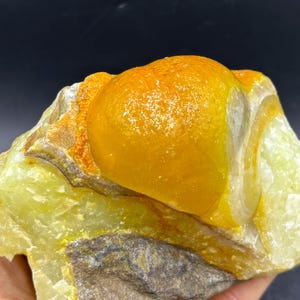 May include: A raw, uncut gemstone featuring a large, rounded, golden-yellow formation atop a base of pale green and gray rock. The stone has a rough, natural texture with areas of orange and white.