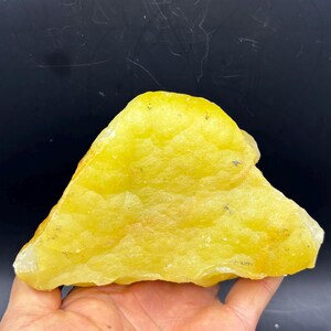 May include: A large, rough-hewn, yellow-colored stone with a triangular shape. The stone has a textured surface and is held in a hand against a dark background. The stone appears to be a natural mineral or crystal specimen.