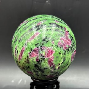 May include: A polished spherical stone featuring a vibrant green base with scattered patches of deep magenta and streaks of black mineral inclusions. The sphere is displayed on a black stand, showcasing a smooth, reflective surface texture.