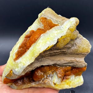 Könnte beinhalten: Natürliches Mineralexemplar mit Schichten in Gelb, Braun und Orange. Der rohe Stein hat eine strukturierte Oberfläche mit kristallinen Formationen. Die Farben sind vielfältig und das Gesamtbild ist natürlich.
