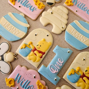 May include: A set of Winnie the Pooh themed sugar cookies decorated with blue, yellow, and pink icing. The cookies feature Winnie the Pooh, a bee, a honeycomb, and the words "Willow" and "Hunny".