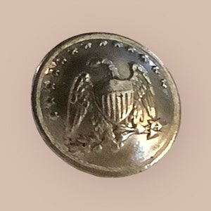 May include: A silver-toned button with an eagle design. The eagle is holding a shield with vertical stripes. The button has a raised rim and a smooth surface.