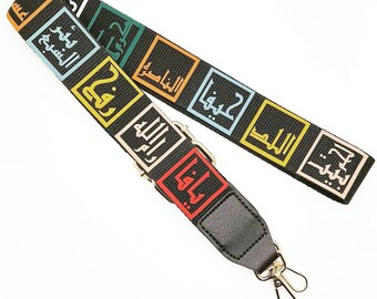 May include: Black and colorful patterned bag strap with Arabic text in various colors. The strap features gold-colored metal hardware and a black leather end piece with a clasp. The text is in square blocks.
