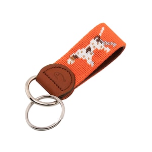 May include: Orange key chain with a brown leather loop and a white and brown dog design. The key chain has two silver key rings.