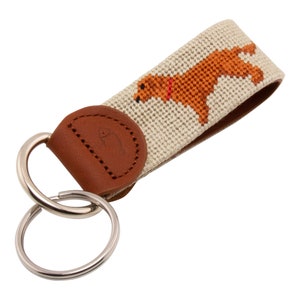 May include: A brown leather key chain with a beige fabric strap featuring a brown dog silhouette. The strap has a white fish logo on the leather. The key chain has two silver key rings.