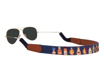 May include: A navy blue sunglass strap with brown leather ends. The strap features a repeating pattern of small, colorful bottles.