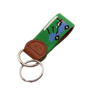 May include: A green and blue key chain with a leather strap and a double ring. The key chain features a small blue car with a person driving it.