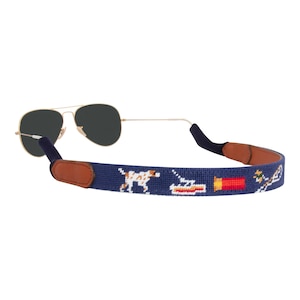 May include: A navy blue sunglass strap with a brown leather end piece. The strap features a white dog, a boat, a shotgun shell, and a bird in flight.