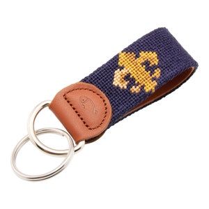 May include: A navy blue key chain with a gold fleur-de-lis design and a brown leather loop with a fish logo. The key chain has two silver key rings.